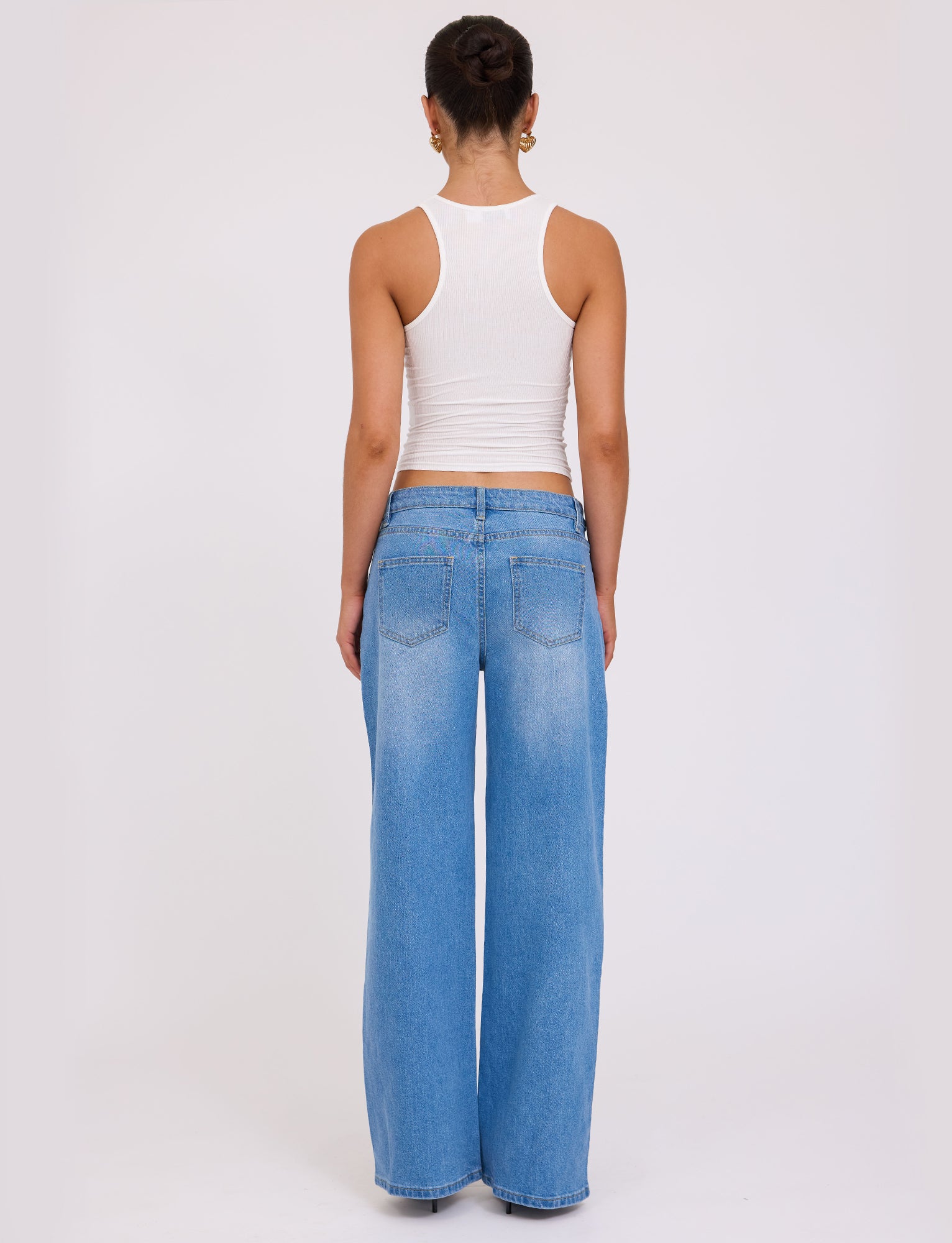 A person with a dark hair bun is standing with their back to the camera, wearing a white sleeveless top and the VEGA JEAN - BLUE : DENIM : MID BLUE from TIGERMIST. The background is plain and white, providing a clean and simple setting.