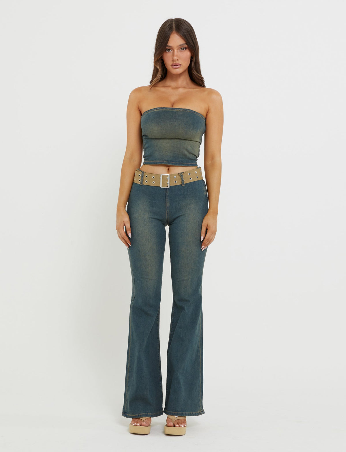 A woman stands against a plain white background, wearing the TIGERMIST ISABEL JEAN - BLUE denim tube top and matching high-waisted, flared jeans. She has long, straight hair and is accessorized with a wide beige belt featuring metallic accents and platform sandals.