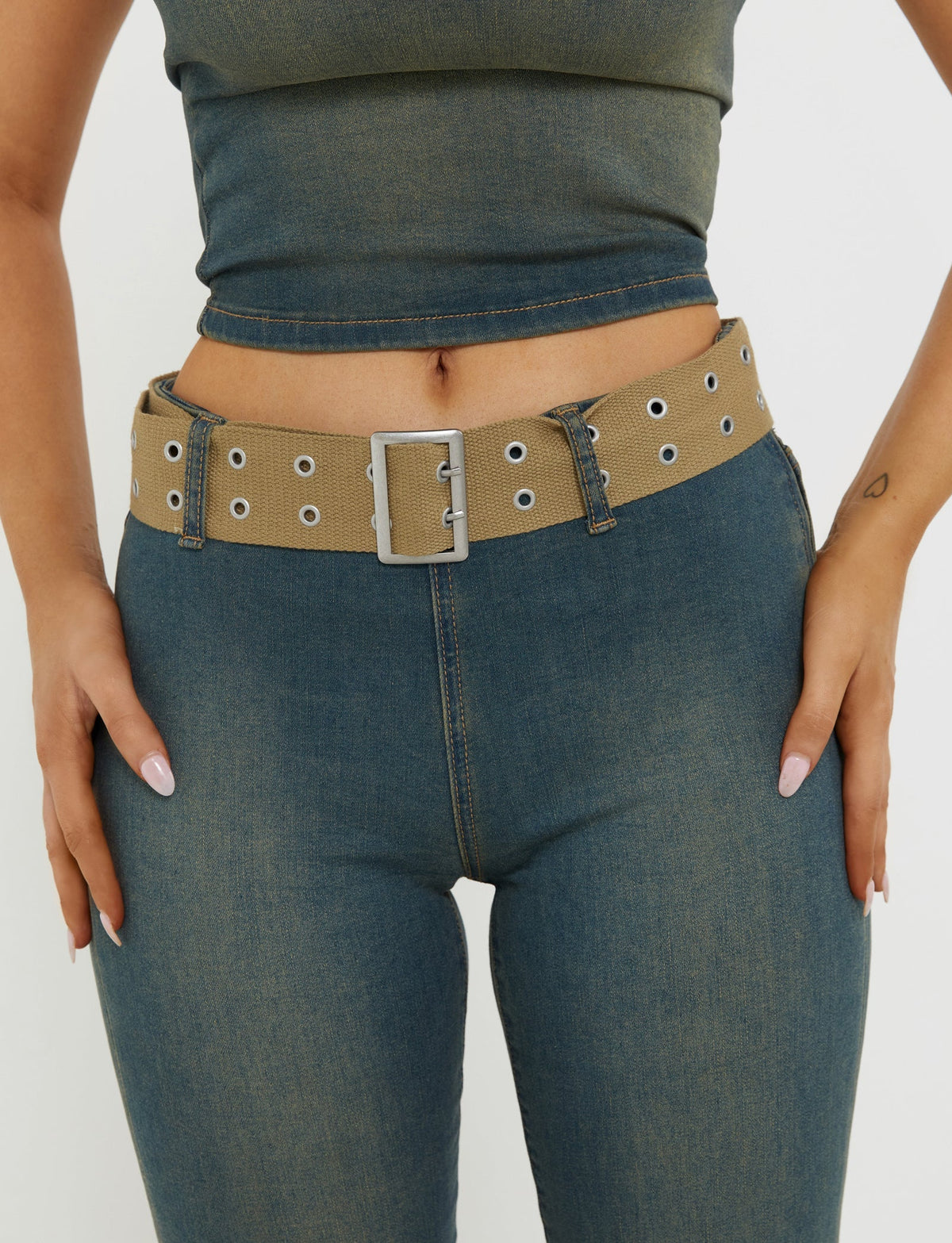 A person is shown from the waist down, dressed in high-waisted ISABEL JEAN - BLUE denim jeans and a matching cropped blue top, both by TIGERMIST. They are wearing a beige belt with silver eyelets and a large rectangular buckle around their waist, with their hands resting on their hips.