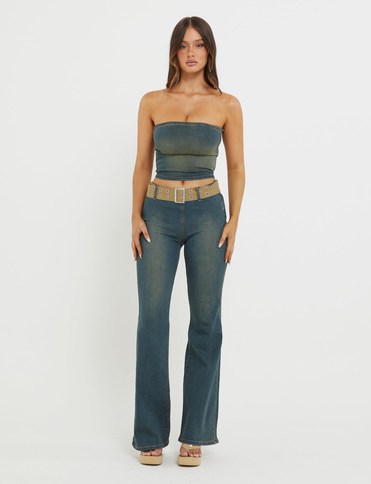 A woman stands against a plain white background, wearing the TIGERMIST ISABEL JEAN - BLUE: DENIM BLUE strapless denim top and high-waisted, flared denim jeans. She accessorizes with a tan belt featuring gold grommets and wooden platform sandals. Her long hair is styled in loose waves, and she faces the camera directly.