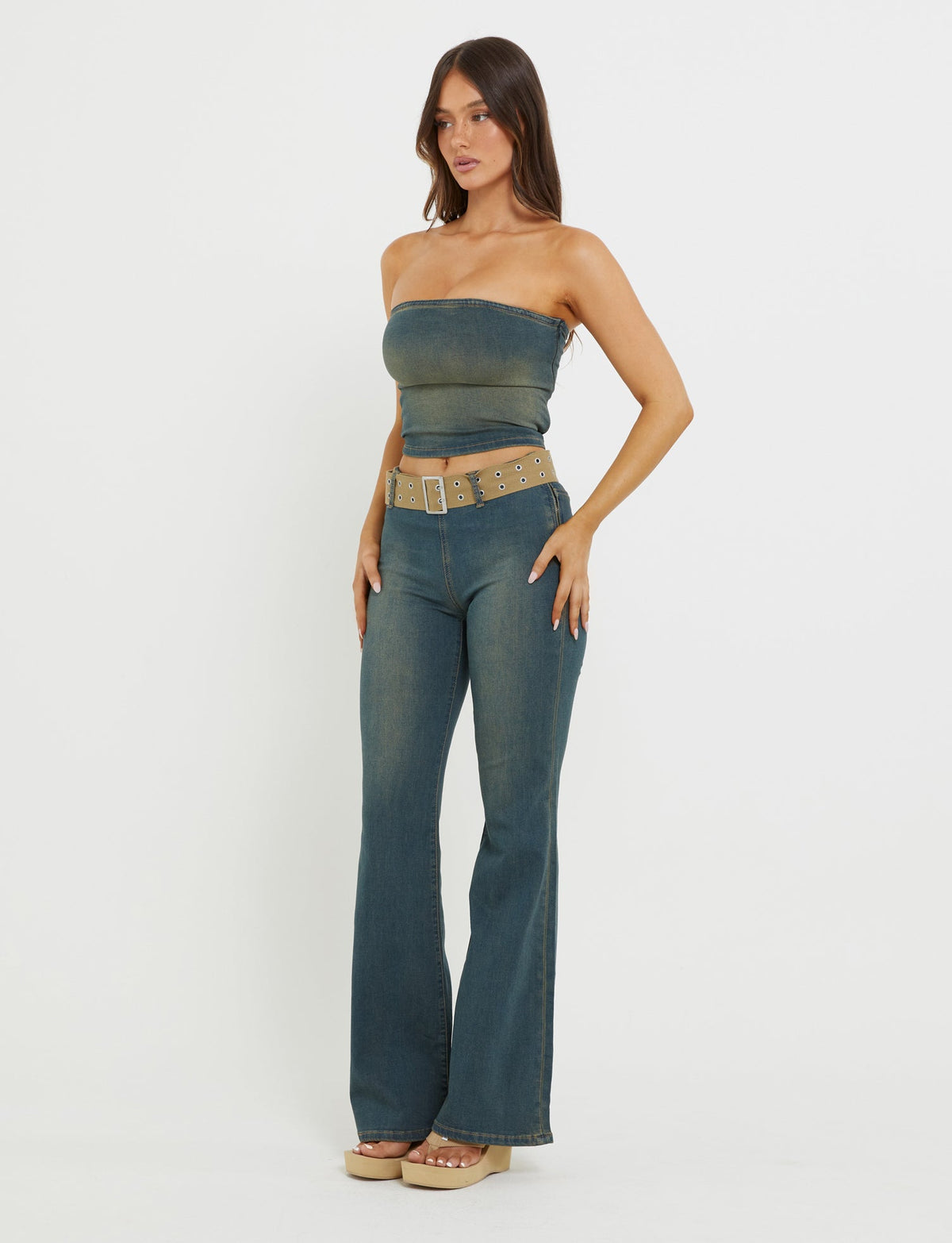 A woman stands against a plain white background, dressed in TIGERMIST's ISABEL JEAN - BLUE: DENIM BLUE with a strapless denim top and high-waisted, flared jeans cinched with a beige belt. She completes her look with platform sandals, her long hair cascading down as she faces slightly to the side with her hands on her hips.