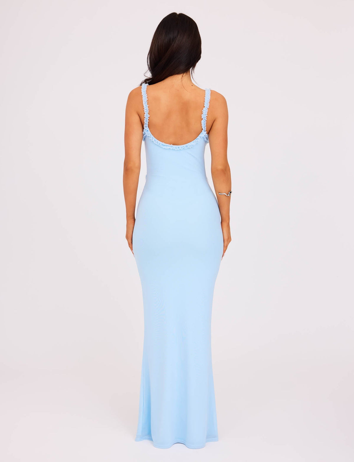 A woman is standing with her back facing the camera, wearing a stunning TIGERMIST RAYLA DRESS in pastel blue. The floor-length dress features thin straps and an open back, perfectly complementing her long dark hair and the bracelet on her right wrist. The background is plain white.