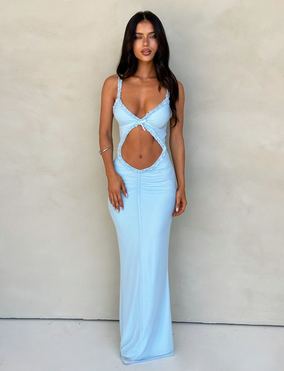 A woman stands against a plain, light-colored background wearing the TigerMist Rayla Dress in pastel blue. This sleeveless, floor-length dress features a striking large diamond-shaped cut-out revealing her midriff. She has long, dark hair and a neutral facial expression and accessorizes with a bracelet on her left wrist.