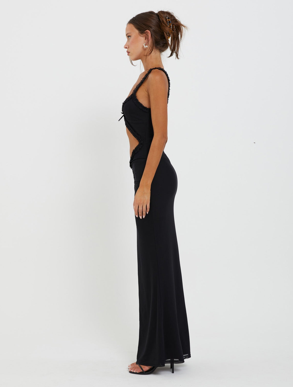 A woman stands in profile against a white background, wearing the RAYLA DRESS - BLACK from TIGERMIST. The form-fitting black dress features cut-out details on the side and a long, flowing skirt. Her hair is styled in an updo, and she accessorizes with earrings and high heels.