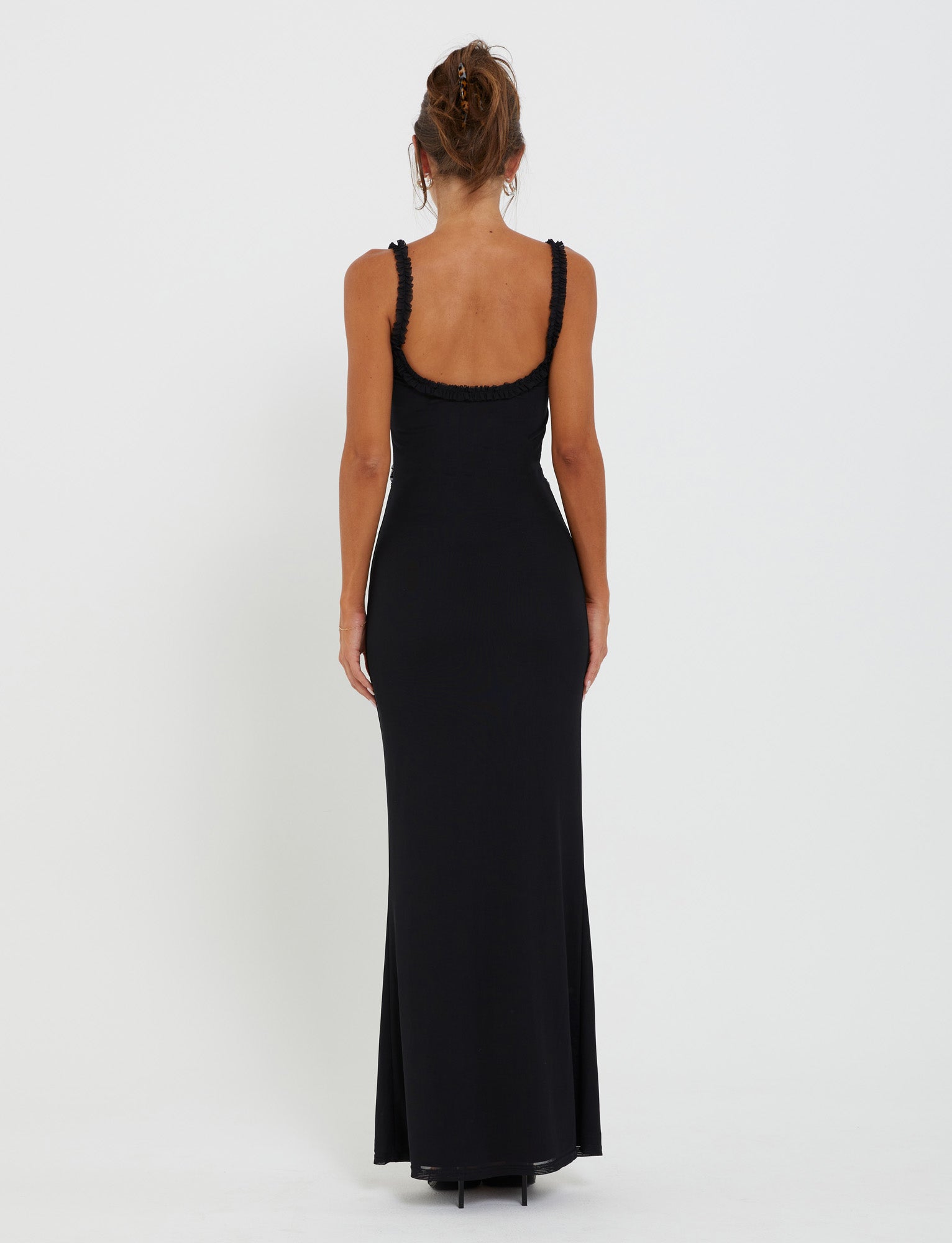 A person with brown hair tied in an updo stands with their back to the camera, wearing a long, sleeveless RAYLA DRESS - BLACK from TIGERMIST, featuring a low-cut back. The background is plain white.