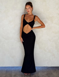 A woman stands against a textured beige wall, wearing a black, form-fitting RAYLA DRESS from TIGERMIST. The dress features a striking cut-out detail at the midriff. Her long hair is pulled back, emphasizing the unique design of the outfit. She accessorizes minimally and pairs her look with black heels.