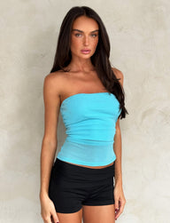 A woman with long brown hair is wearing a bright blue sparkle TIGERMIST HARRIETT TUBE TOP and black shorts. She is standing against a plain light-colored wall, looking directly at the camera with a neutral expression.