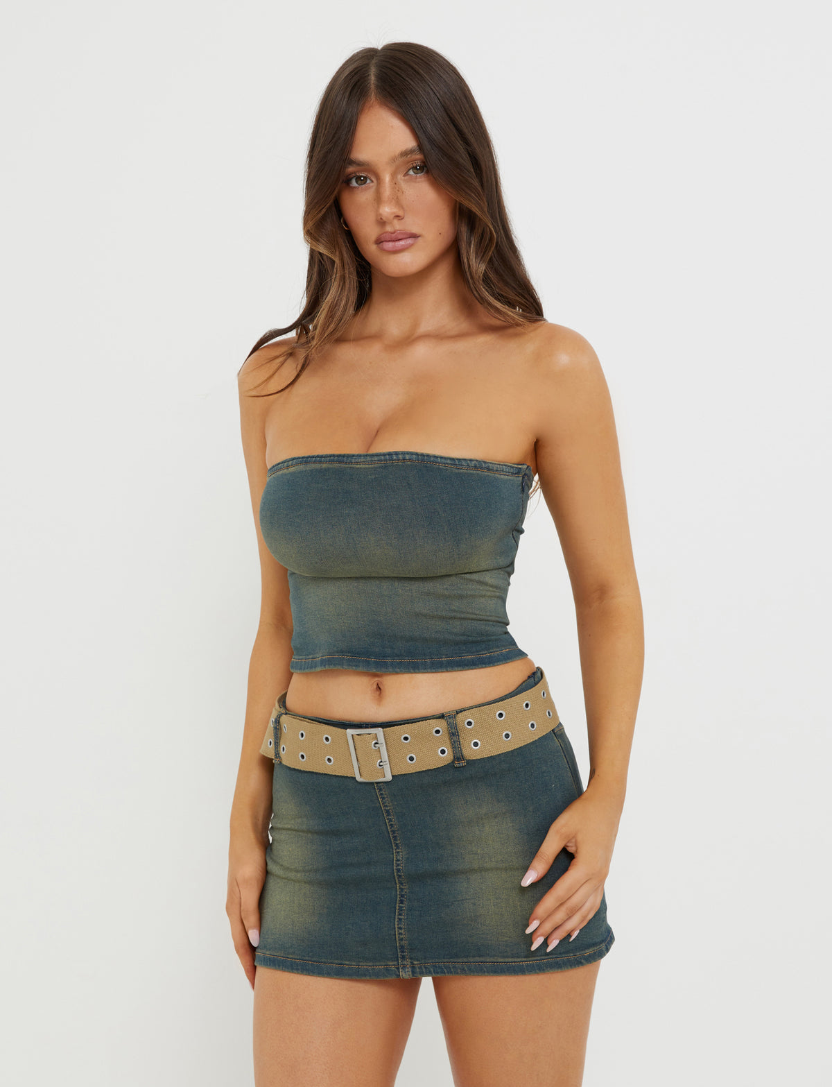 A woman with long brunette hair is wearing the ISABEL SKORT - BLUE from TIGERMIST. She pairs the denim blue skort with a matching strapless, denim tube top and a tan belt. She is standing against a plain white background.