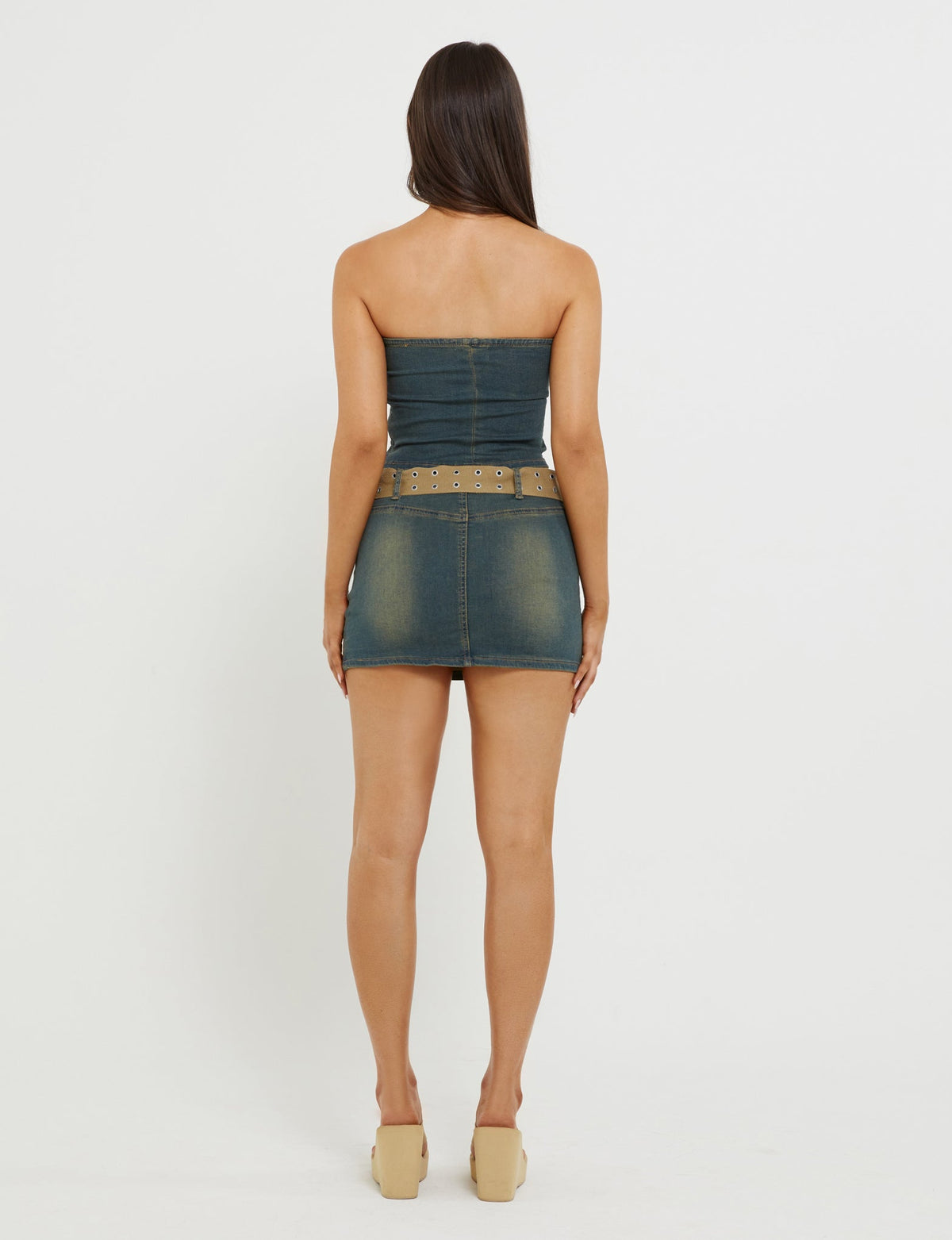 A person with long brown hair stands facing away from the camera in a fitted TigerMist ISABEL SKORT - BLUE, ending above the knees, paired with a matching belt with metal eyelets and tan platform shoes. The background is plain white.