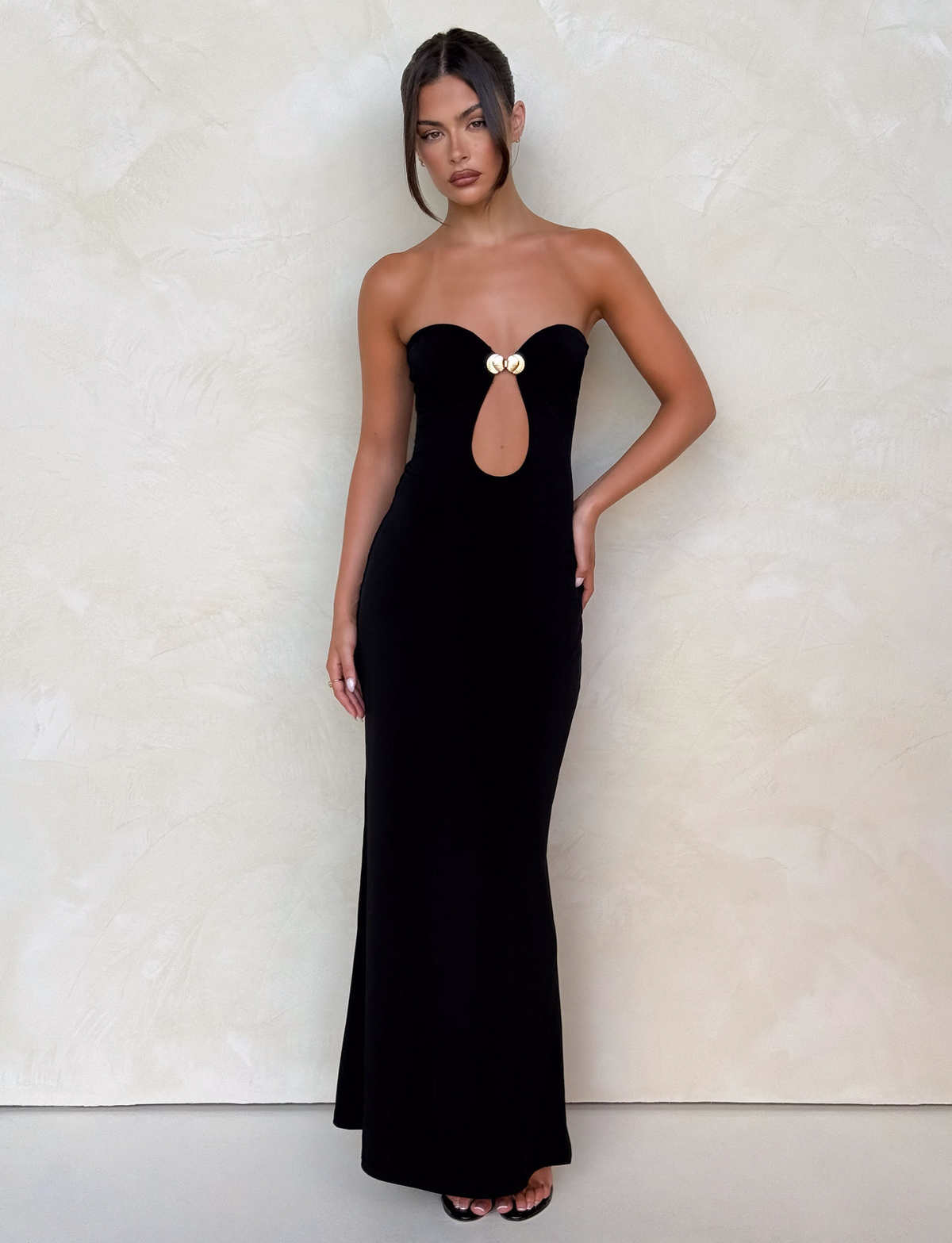 A woman poses confidently against a light textured wall, wearing the TIGERMIST CECILY MAXI DRESS - BLACK—a chic strapless maxi with keyhole cutout and gold neckline accent. Her hair is styled up, and she places one hand on her hip.