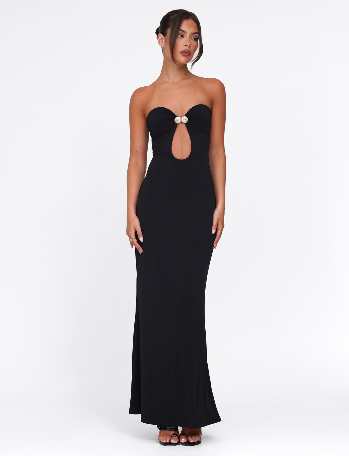 A woman poses in the TIGERMIST CECILY MAXI DRESS - BLACK, a strapless, floor-length gown with a sweetheart neckline, front cutout, and embellished bust. She completes the look with black heels and an updo against a white background.