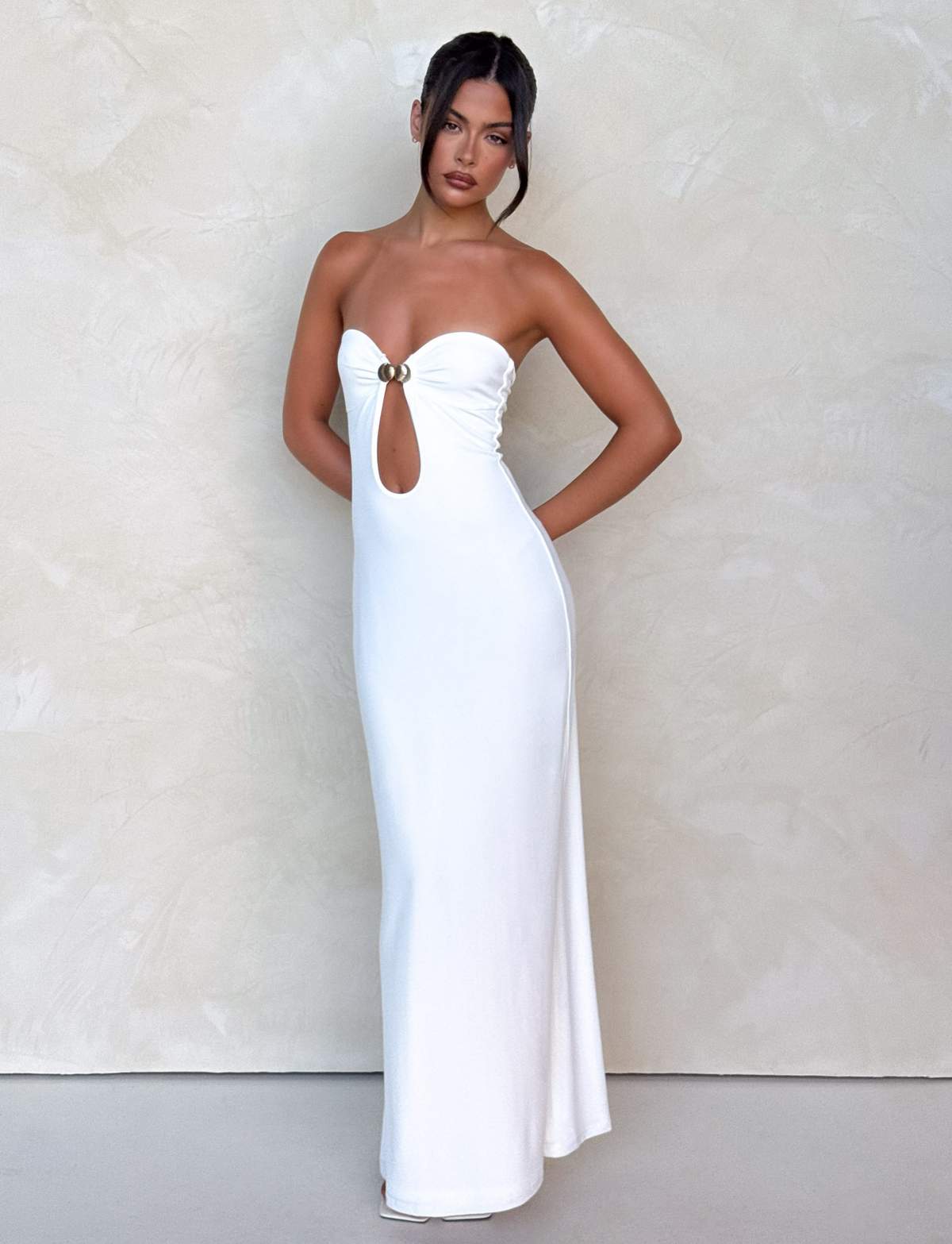 A woman models the TIGERMIST CECILY MAXI DRESS in white, a strapless, fitted, floor-length dress with a front cutout and gold ring detail, posing against a light textured background with her arms behind her.