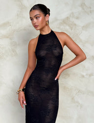 A person stands against a textured beige wall, wearing the exquisite LUCIA DRESS - BLACK : LACE from TIGERMIST. They have an elegant hairstyle, gold hoop earrings, and several gold bracelets on their left wrist. Their right hand rests on their hip as they look off to the left with a neutral expression.