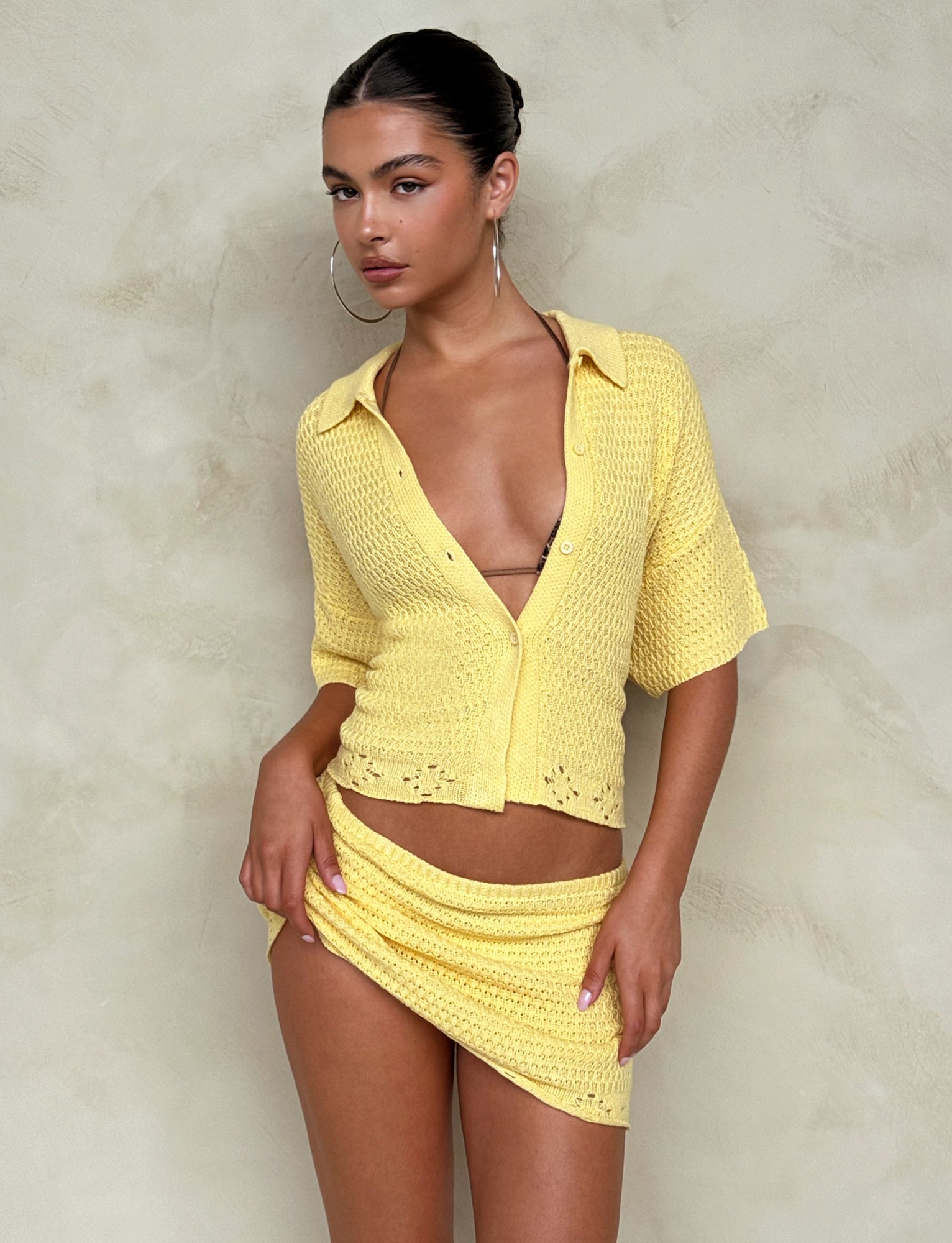 A person stands posing against a textured neutral background, donning Tiger Mist's JANAI SKIRT - YELLOW : LEMON, a high-waisted mini skirt with a matching yellow knitted short-sleeve top featuring a deep neckline. Large hoop earrings and natural makeup complete the look, conveying a stylish and confident appearance.