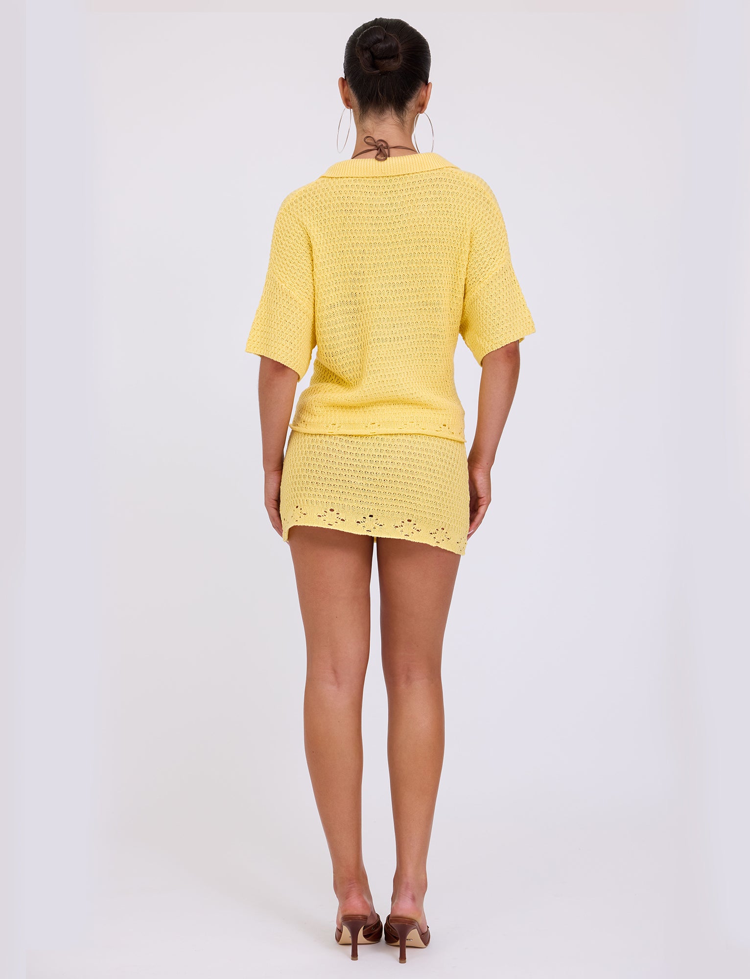 A woman is standing with her back to the camera, wearing a short yellow JANAI TOP from TigerMist and high-heeled sandals. Her hair is styled in a bun, and she is adorned with large hoop earrings. The background is plain white.