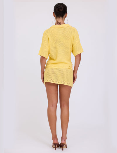 A woman is standing with her back to the camera, wearing a short yellow JANAI TOP from TigerMist and high-heeled sandals. Her hair is styled in a bun, and she is adorned with large hoop earrings. The background is plain white.