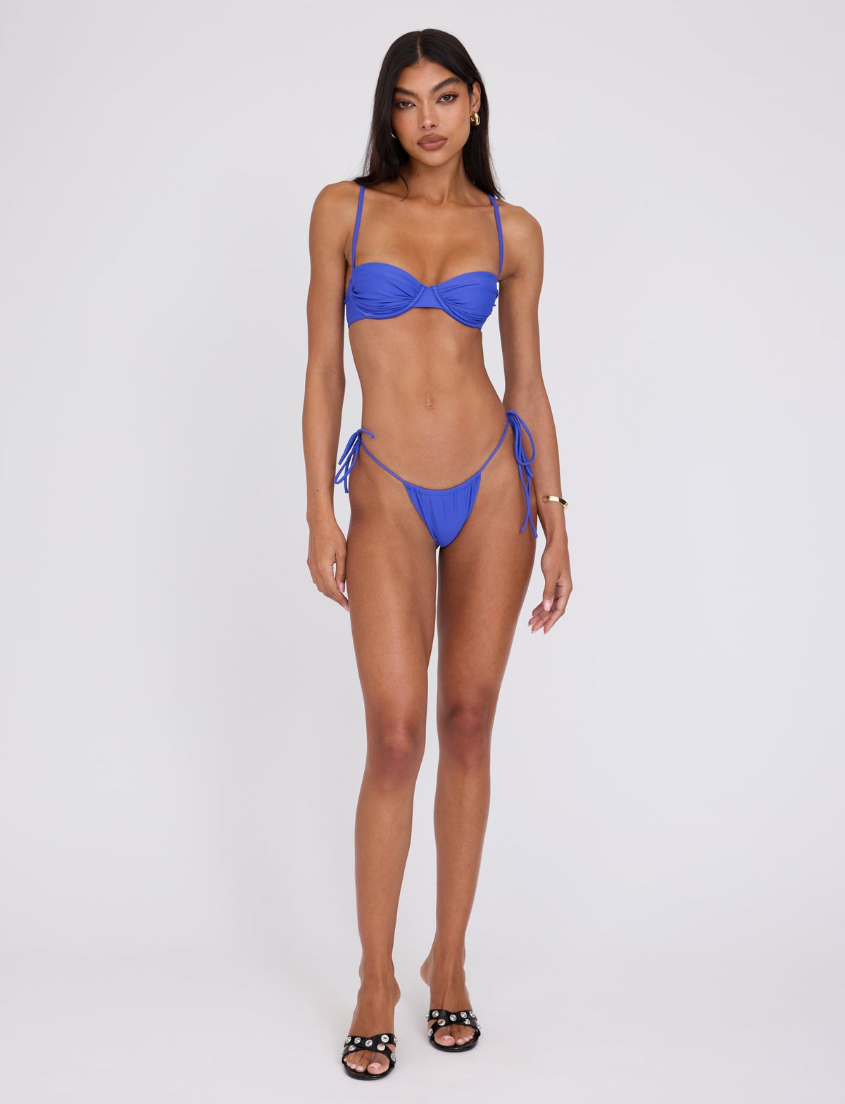 A woman stands against a plain white background, wearing the TIGERMIST KHESSA BIKINI TOP in cobalt blue. She has long dark hair and is looking directly at the camera. She is also wearing black sandals and a gold bracelet on her left wrist.