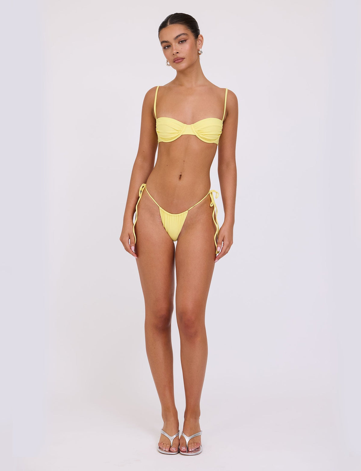 A woman stands against a plain white background wearing the KHESSA BIKINI TOP - YELLOW : LEMON from TIGERMIST. The bikini top features thin straps and a ruched design, paired with bottoms tied at the sides. She wears flip-flop sandals and has her hair pulled back. She looks straight at the camera.
