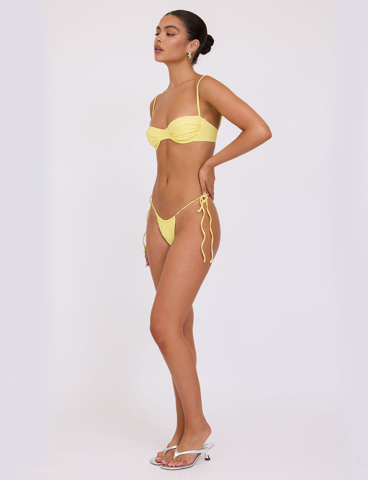 A woman is posing against a white background, wearing the yellow TIGERMIST KHESSA BIKINI TOP - LEMON with side ties and white sandals. She has her hair tied back and stands in a relaxed pose, looking to the left.