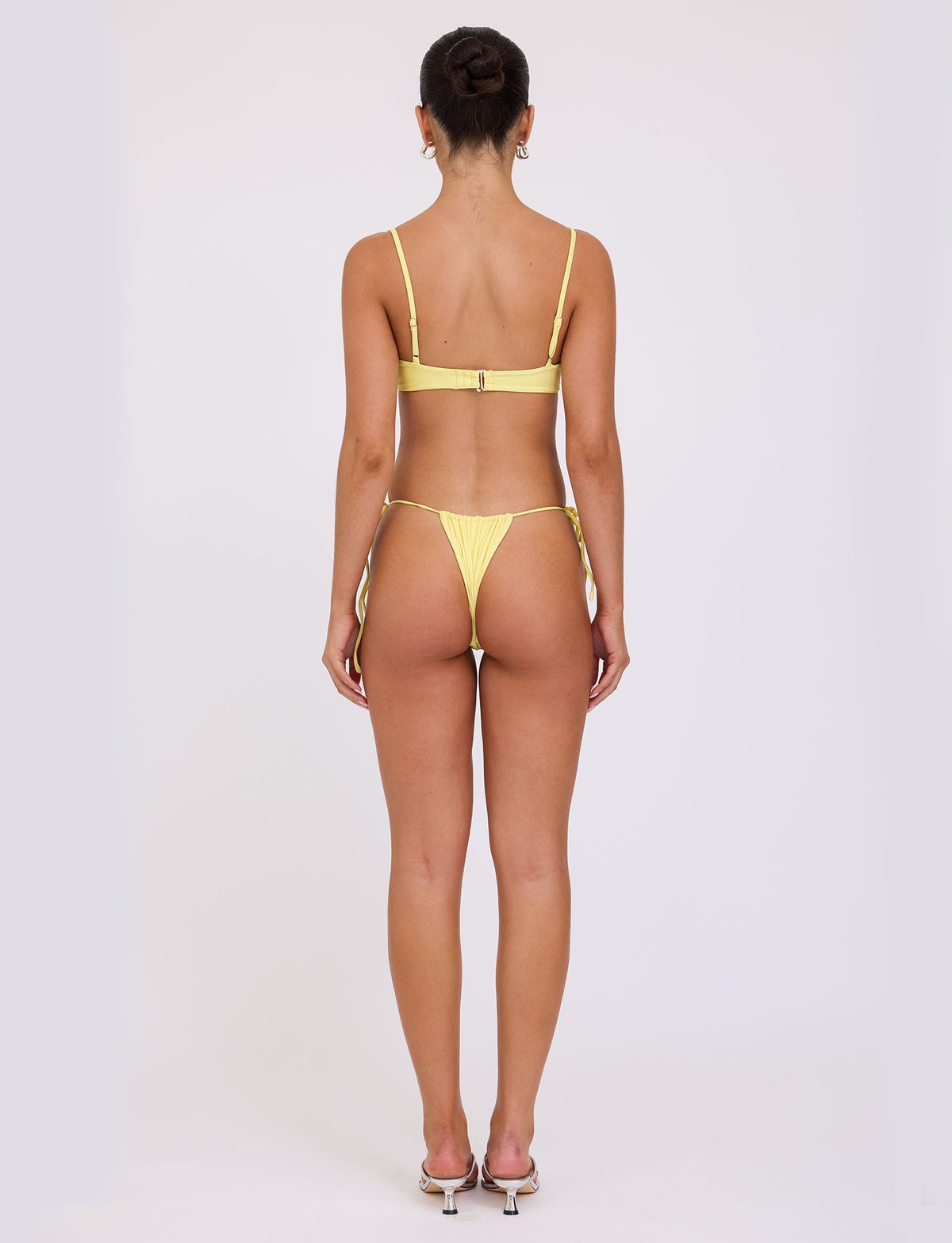 A person wearing the KHESSA BIKINI TOP - YELLOW : LEMON from TIGERMIST stands facing away from the camera. The bikini has spaghetti straps and ties on the sides of the bottoms. They are standing on a plain white background and wearing high-heeled shoes.