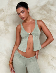 A woman stands against a textured beige wall, wearing the Tiger Mist AMETHYST TOP - GREEN in green sparkle, which features a deep V-neck fastened with a small metal clasp. She pairs it with matching high-waisted pants. Her hair is styled in a sleek updo, and she accessorizes with gold hoop earrings and a bracelet.