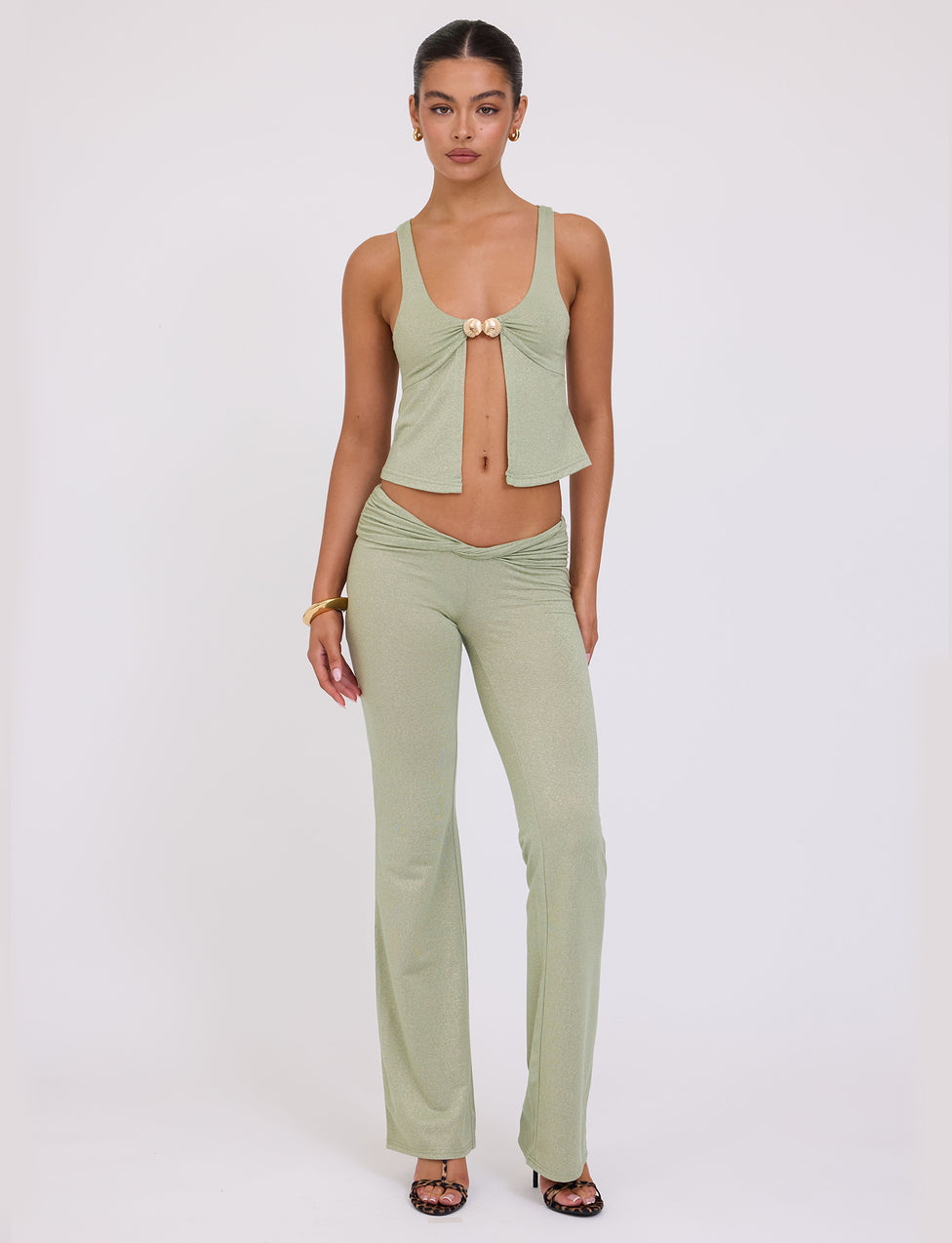 A woman stands against a plain white background, wearing the AMETHYST PANT - GREEN SPARKLE from TIGERMIST. The top features a deep neckline and front tie detail, while the high-waisted pants are flared. She accessorizes with a gold bracelet and black heeled sandals.
