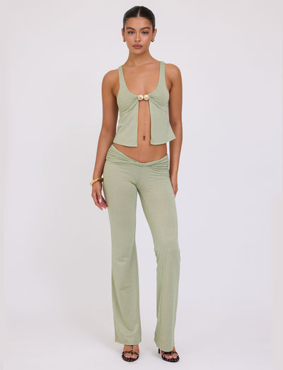 A person stands against a white backdrop wearing the LIGHT GREEN SPARKLE "AMETHYST" SLEEVELESS TOP by TIGERMIST, featuring a front tie at the bust, paired with matching high-waisted flared pants. They accessorize with hoop earrings, a bracelet, and open-toe heels. Their expression is neutral and their hairstyle is sleek.