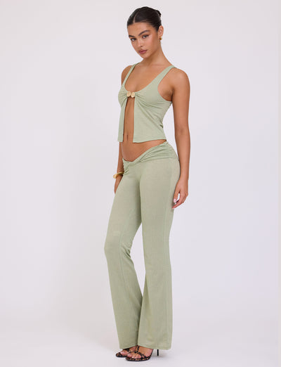 A woman stands against a plain white background, wearing the chic green sparkle Amethyst Top by TigerMist. Her stylish ensemble includes a sleeveless top with a front tie and high-waisted, flared pants. With her hair pulled back and black strappy heels, she effortlessly exudes chic elegance.