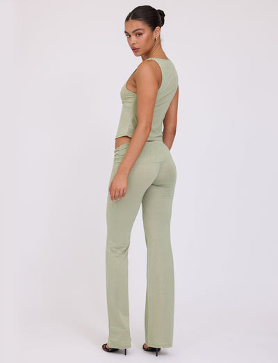 A woman with dark hair in a sleek bun is wearing the AMETHYST TOP - GREEN : GREEN SPARKLE from TIGERMIST along with matching high-waisted pants. She is standing sideways, turning her head slightly to look over her shoulder, against a plain white background.