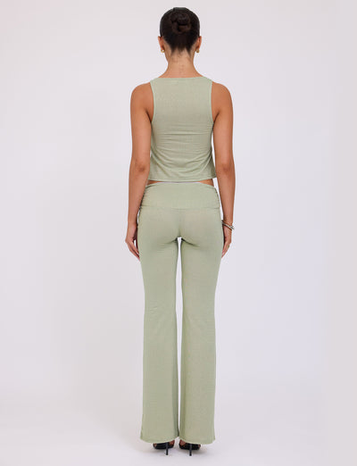 A person is seen from the back, standing with their hair in a bun. They are wearing a light green sleeveless top and matching AMETHYST PANT - GREEN : GREEN SPARKLE from TIGERMIST, along with black heels. The background is a plain, light-colored surface.