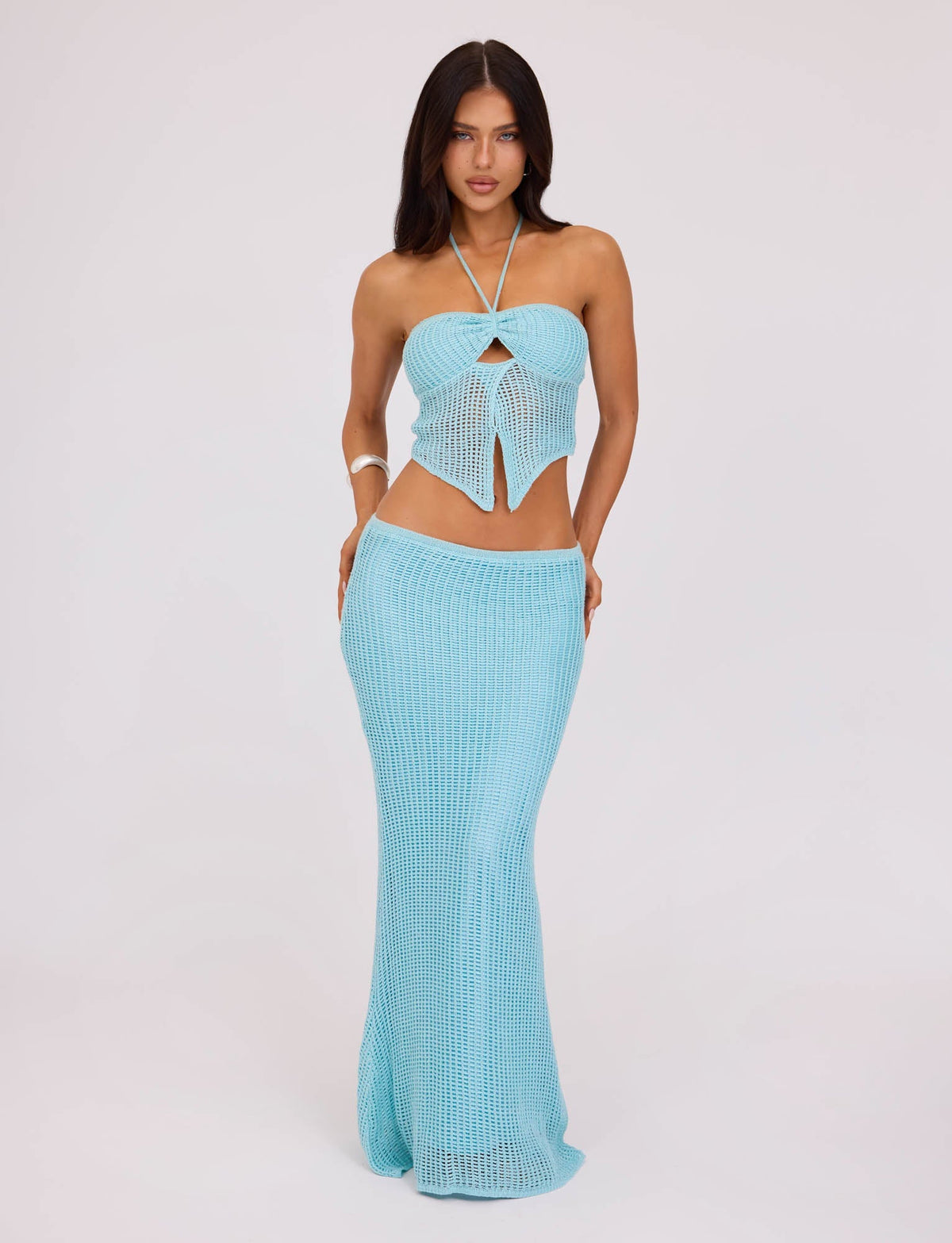 A woman stands against a plain background wearing the TIGERMIST SHAH SKIRT in pastel blue, a crochet-style two-piece outfit that includes a halter top with a keyhole cutout and a long, fitted skirt. She has long, dark hair and a neutral expression.