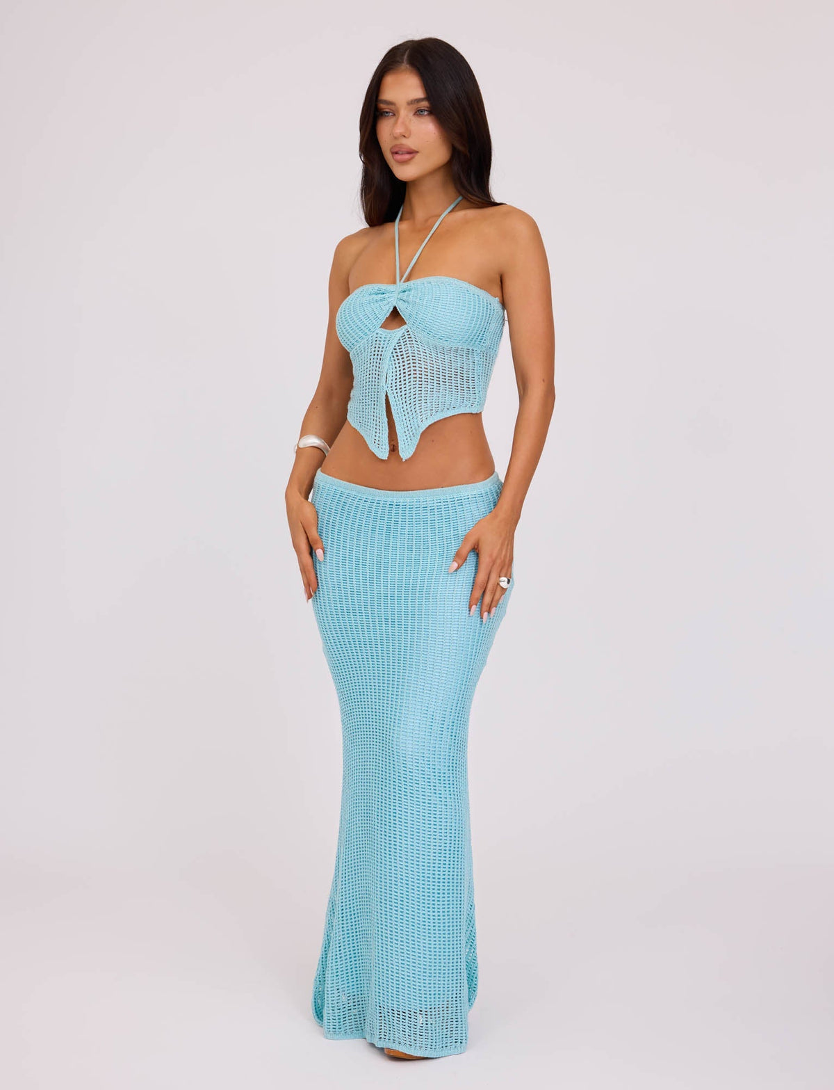 A woman stands against a plain background wearing a pastel blue crochet halter-top and the SHAH SKIRT in pale blue from TIGERMIST. Her long, dark hair cascades over her shoulders as she rests her hands lightly on her hips, exuding confidence and style.