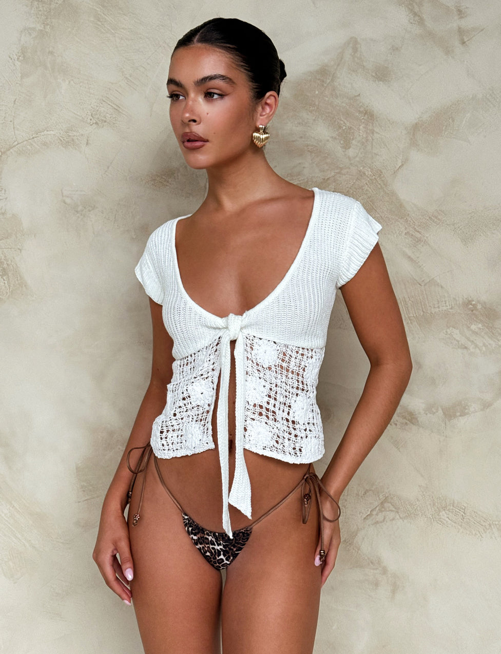 A woman with her hair tied back is posing against a beige textured wall. She is wearing the TAYA TOP - WHITE from TIGERMIST, a short-sleeved crochet top tied in the front, paired with a Tiger Mist leopard-print bikini bottom. Her look is completed with large gold hoop earrings.