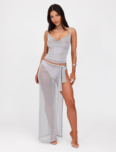 A woman stands against a white background wearing the WYNNE TOP - GREY : SILVER from TIGERMIST. The sheer, light gray two-piece outfit features a silver metallic knit. The fitted silhouette includes a camisole top with adjustable shoulder straps and a wraparound skirt with a high slit, revealing her legs. She has long dark hair and is wearing high-heeled sandals.