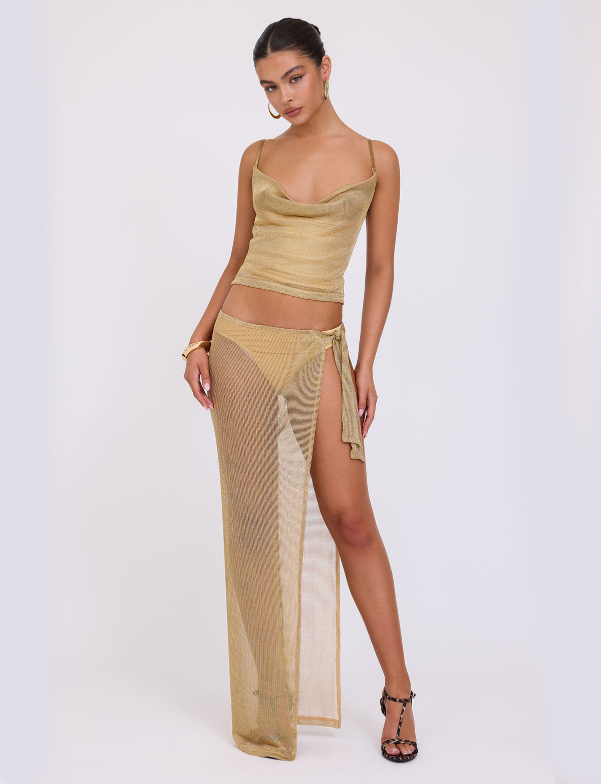 A person stands in a confident pose wearing the gold, semi-sheer WYNNE TOP from TIGERMIST. It is paired with a high-slit skirt over a bikini bottom. They have dark hair pulled back, accessorizing with hoop earrings and black heels.