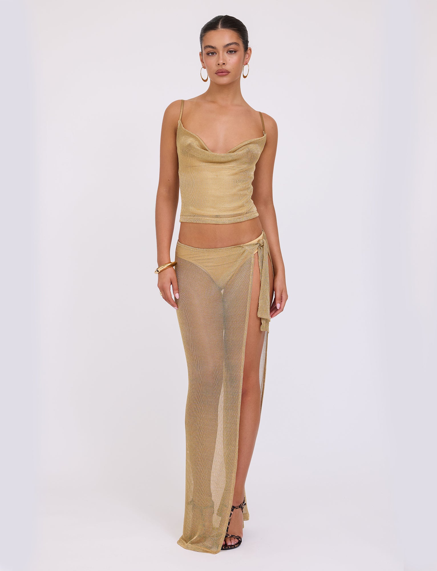 A woman stands against a plain white background wearing the WYNNE TOP - GOLD from TIGERMIST, paired with a matching semi-transparent golden long skirt featuring a side slit. She accessorizes with hoop earrings, a bracelet, and black strappy heels.