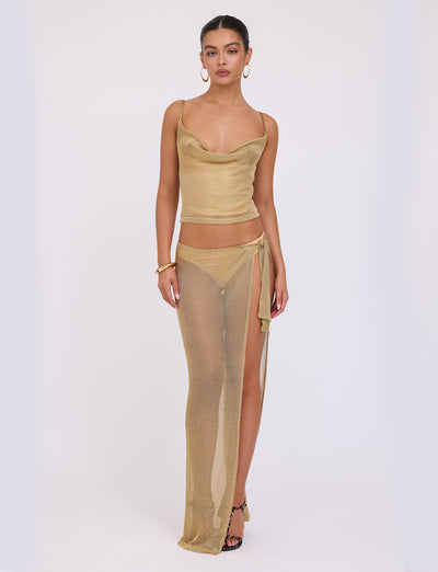 A woman stands against a plain white background wearing the WYNNE TOP - GOLD from TIGERMIST, paired with a matching semi-transparent golden long skirt featuring a side slit. She accessorizes with hoop earrings, a bracelet, and black strappy heels.