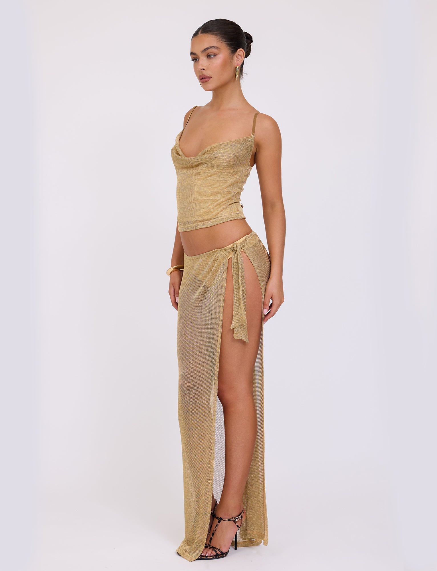 A woman poses against a plain white background, dressed in the TIGERMIST WYNNE TOP - GOLD, a semi-sheer two-piece outfit. The top features thin straps and a draped neckline, complemented by a skirt with a high slit tied at the hip. She completes her look with high-heeled sandals and has her hair pulled back in a bun.