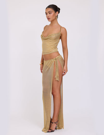 A woman poses against a plain white background, dressed in the TIGERMIST WYNNE TOP - GOLD, a semi-sheer two-piece outfit. The top features thin straps and a draped neckline, complemented by a skirt with a high slit tied at the hip. She completes her look with high-heeled sandals and has her hair pulled back in a bun.