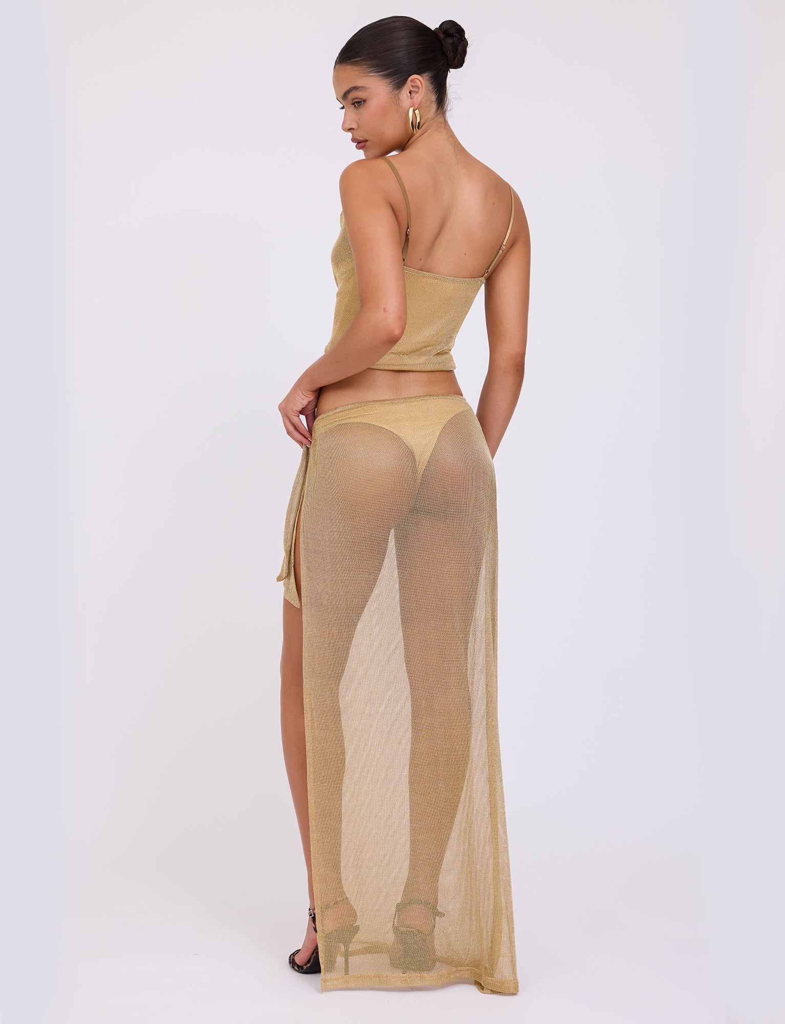 A woman stands with her back to the camera, wearing the gold WYNNE TOP - GOLD from TIGERMIST, paired with a matching sheer long skirt. Her hair is styled in a sleek bun, and she accessorizes with hoop earrings and high heels.