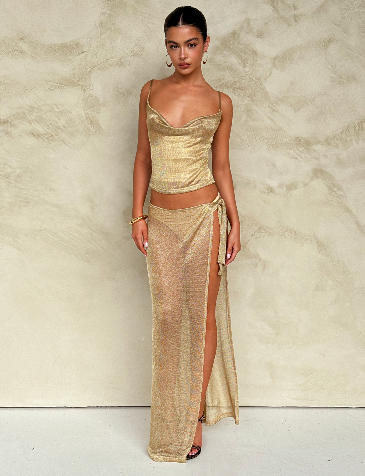 A woman stands against a textured beige wall, wearing the TIGERMIST WYNNE SKIRT in gold with a matching sleeveless top. She accessorizes with hoop earrings, a bracelet, and black heels, her hair slicked back and expression neutral.