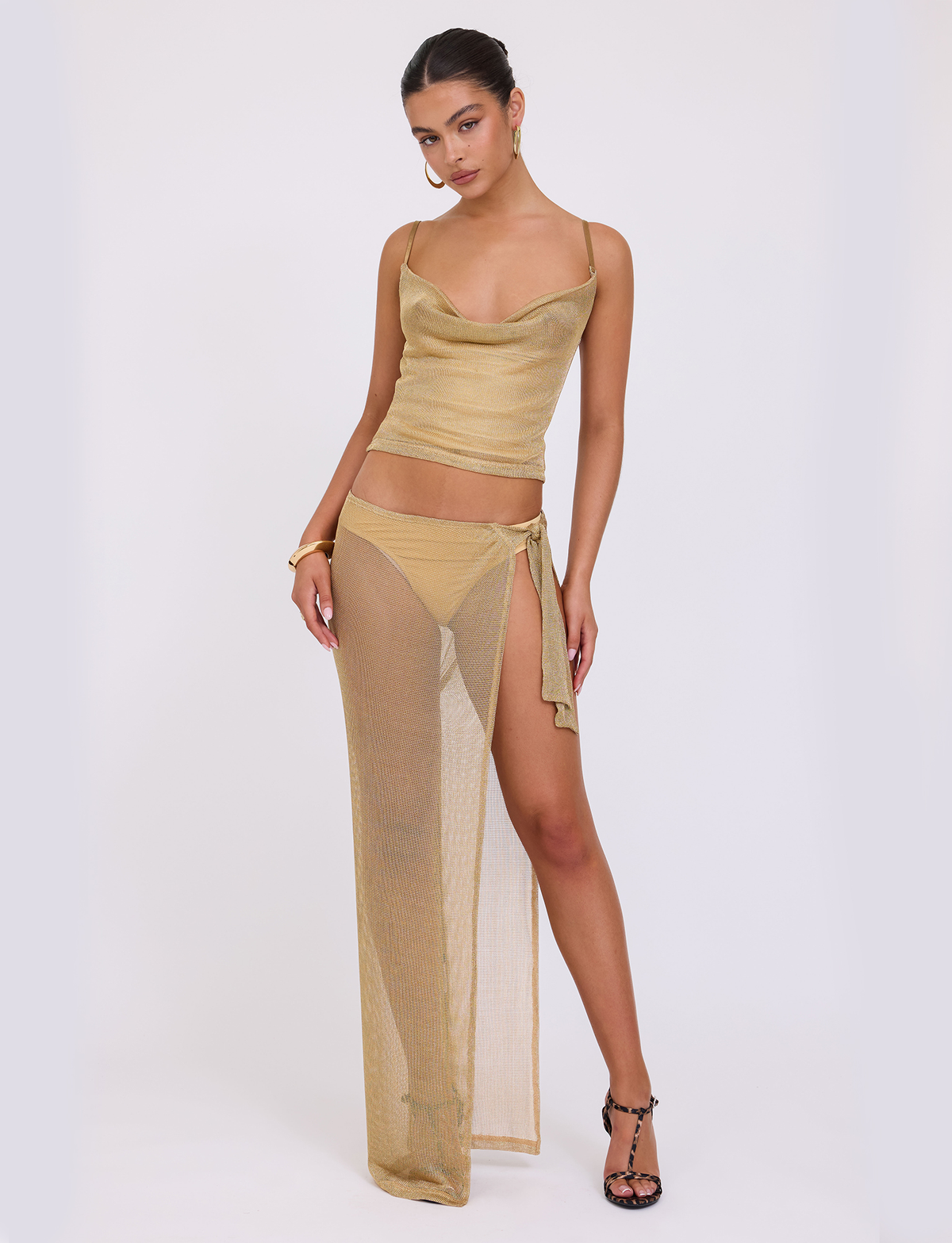 A woman models TIGERMIST's WYNNE SKIRT in gold—a semi-sheer metallic knit maxi with a high slit—paired with a cowl-neck camisole, high-waisted bikini bottoms, hoop earrings, and strappy black heels.