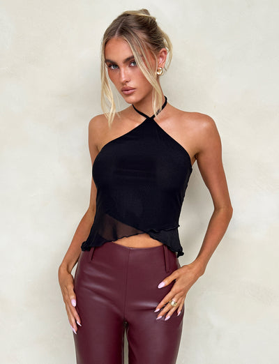 A woman with blonde hair styled in a loose updo wears the HESTIA TOP - BLACK, a sleek mesh halter top, and high-waisted burgundy leather pants, showcasing a fitted silhouette. She accessorizes with hoop earrings and several rings on her fingers. Standing against a neutral, pale background, she rests her hands on her thighs.
