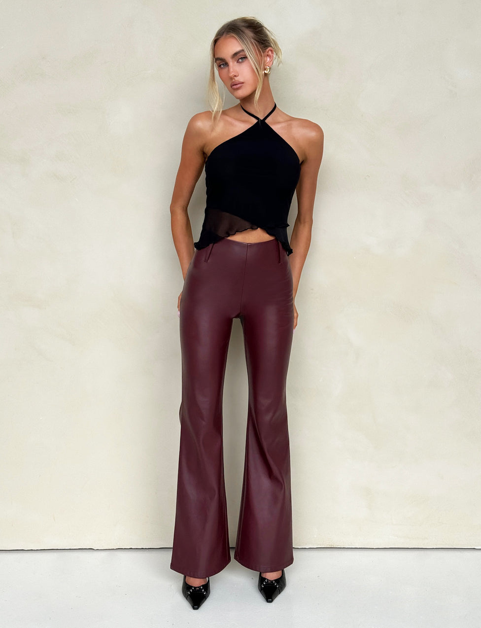 A woman with blonde hair styled in a loose updo wears the HESTIA TOP - BLACK, a sleek mesh halter top, and high-waisted burgundy leather pants, showcasing a fitted silhouette. She accessorizes with hoop earrings and several rings on her fingers. Standing against a neutral, pale background, she rests her hands on her thighs.
