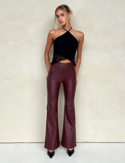A woman stands against a light beige wall, wearing a black halter top and high-waisted, cherry red leather pants from Tiger Mist. Her hair is styled in a loose updo, with a few strands framing her face. She completes the look with black patent leather pointed-toe shoes.