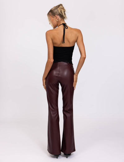 A woman with blonde hair styled in a loose updo wears the HESTIA TOP - BLACK, a sleek mesh halter top, and high-waisted burgundy leather pants, showcasing a fitted silhouette. She accessorizes with hoop earrings and several rings on her fingers. Standing against a neutral, pale background, she rests her hands on her thighs.
