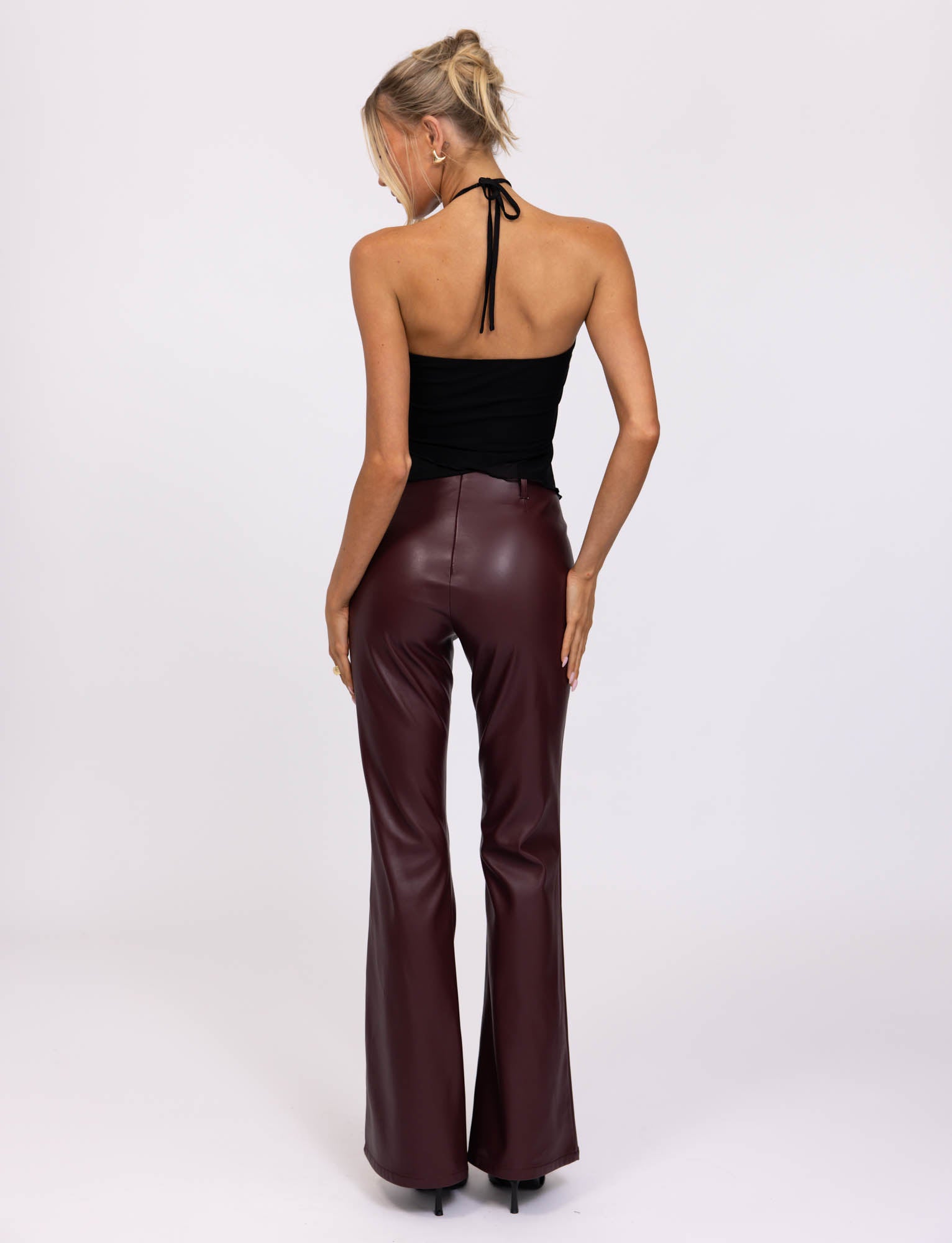 A blonde-haired woman is wearing a sleeveless, black halter top and the high-waisted PU TEDDIE PANT in Cherry Red faux leather with flared legs from TIGERMIST. She stands facing away from the camera against a plain white background.