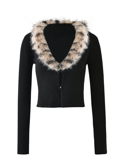 The DARJA CARDIGAN - BLACK from TIGERMIST is a black long-sleeved crop top featuring a deep V-neck collar. It is adorned with a thick, fluffy faux fur trim in a white and black spotted pattern and has a single button closure at the front.