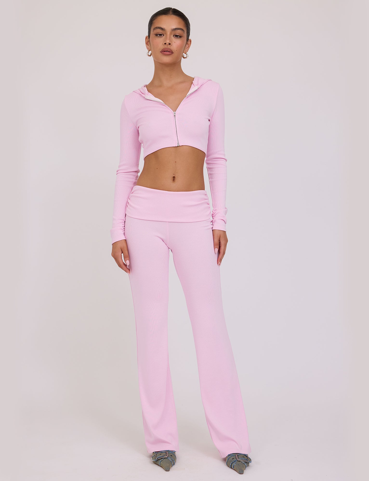 A woman stands against a plain background, wearing a stylish TIGERMIST KARINA JUMPER in light pink, which includes a fitted hooded crop top and matching high-waisted flare pants. She pairs the ensemble with gray shoes, and her hair is pulled back.