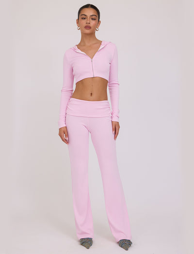 A woman stands against a plain background, wearing a stylish TIGERMIST KARINA JUMPER in light pink, which includes a fitted hooded crop top and matching high-waisted flare pants. She pairs the ensemble with gray shoes, and her hair is pulled back.